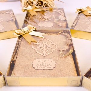 May include: A set of gold gift boxes with a gold prayer rug, a gold prayer bead necklace, and a gold charm. The boxes are decorated with a gold bow and a gold tag that says "Ramadan Mubarak Love Abdul, Anisha & Family xxx".