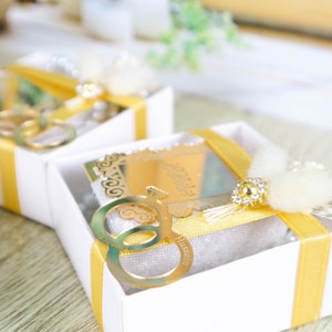 May include: Two white gift boxes with yellow ribbon and a gold key charm. The key charm has two interlocking circles and the name "Hussain" engraved on it. The boxes are decorated with white feathers.