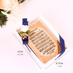 May include: A small wooden box with a blue ribbon and a white flower. The box contains a wooden plaque with Arabic calligraphy and the text "Ayatul Kursi" and "Amir & Zainab 12.12.2022". The box measures 4.1 inches by 2.75 inches.