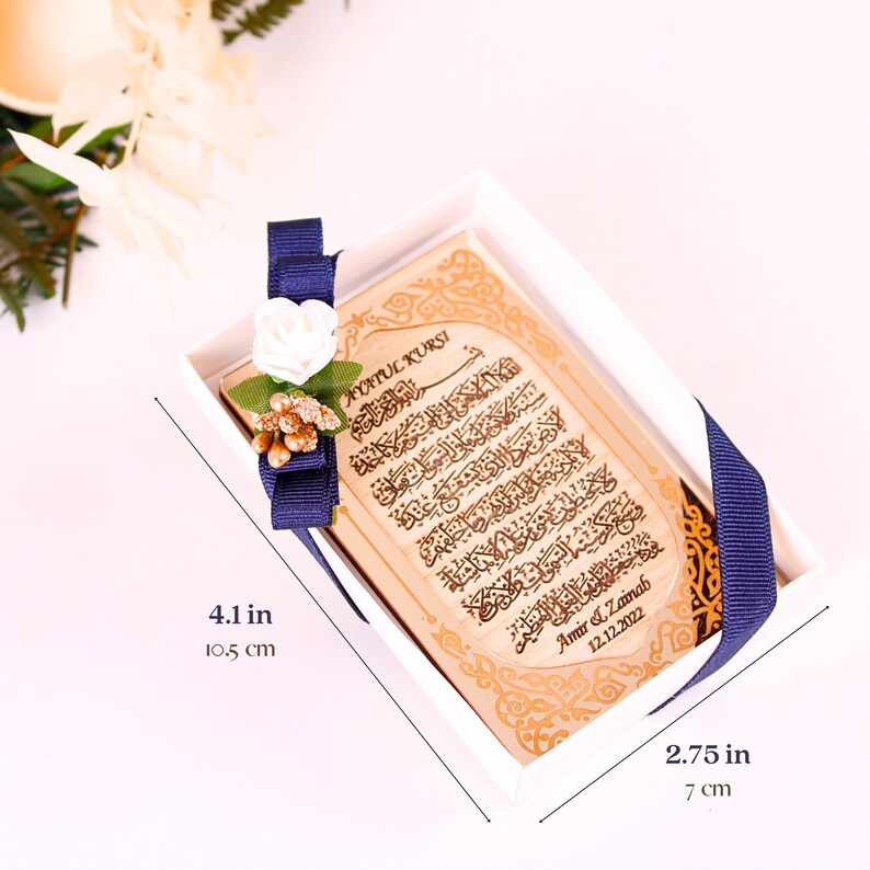 May include: A small wooden box with a blue ribbon and a white flower. The box contains a wooden plaque with Arabic calligraphy and the text "Ayatul Kursi" and "Amir & Zainab 12.12.2022". The box measures 4.1 inches by 2.75 inches.
