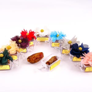 Könnte beinhalten: Sortiment aus durchsichtigen Plastikboxen, die mit bunten Stoffblumen verziert sind. Jede Box enthält eine Dattel und hat ein goldenes Etikett mit dem Text "Ramadan Kareem".