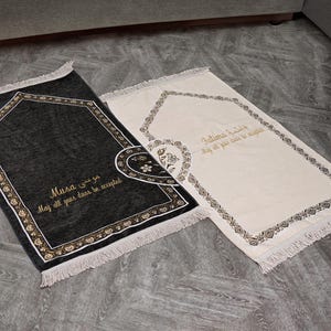 Puede incluir: Dos alfombras de oración, una negra y otra crema, con bordes decorativos y flecos. La alfombra negra tiene el nombre "Musa" y la alfombra crema tiene el nombre "Fatima" bordados en dorado, junto con una oración en inglés.