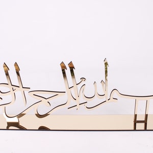 May include: Gold-colored metal wall decor with Arabic calligraphy that reads "Bismillah Al Rahman Al Rahim".