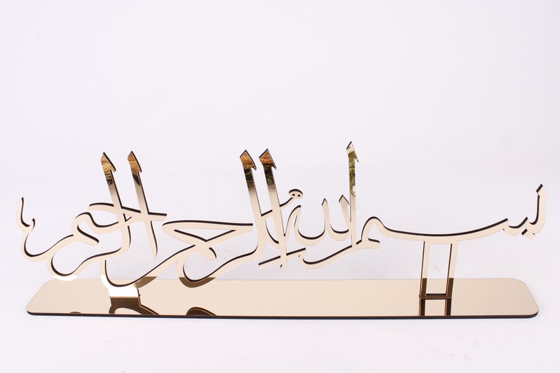 May include: Gold-colored metal wall decor with Arabic calligraphy that reads "Bismillah Al Rahman Al Rahim".