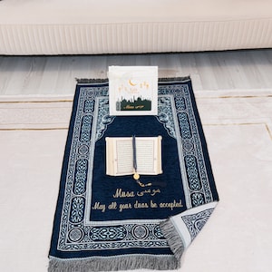 May include: A navy blue prayer mat with a decorative border and the text "Musa" in English and Arabic. An open book, prayer beads, and a greeting card are on the mat. The mat is on a light-coloured rug.