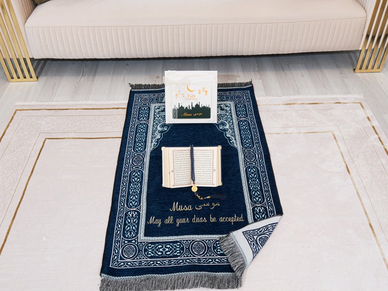 May include: A navy blue prayer mat with a decorative border and the text "Musa" in English and Arabic. An open book, prayer beads, and a greeting card are on the mat. The mat is on a light-coloured rug.