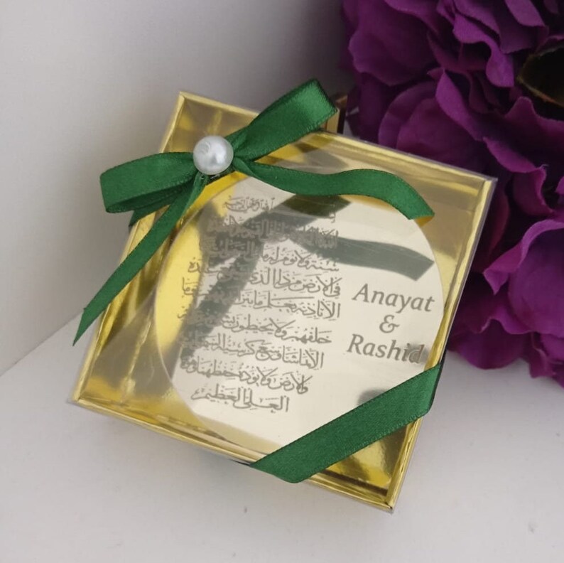 Op de afbeelding: Een ronde gouden doos met een groen lint en een parel accent. De doos bevat een ronde gouden plaquette met Arabische kalligrafie en de gegraveerde namen "Anayat & Rashid".
