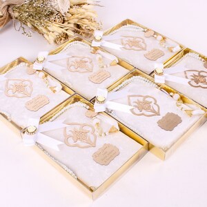May include: Six gift boxes with white prayer mats, pearl prayer beads, and wooden embellishments. Each box is tied with a white ribbon and a gold-colored decorative element. A bouquet of dried flowers is in the background.