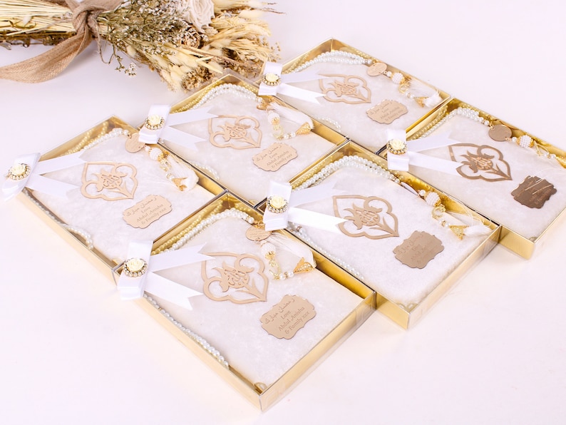 May include: Six gift boxes with white prayer mats, pearl prayer beads, and wooden embellishments. Each box is tied with a white ribbon and a gold-colored decorative element. A bouquet of dried flowers is in the background.