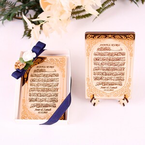 May include: A wooden box with a blue ribbon and a white flower. Inside the box is a wooden plaque with the text "Ayatul Kursi" and Arabic calligraphy. The plaque is engraved with the names "Amir & Zainab" and the date "12.12.2022".