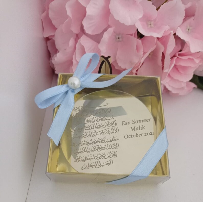 Op de afbeelding: Een klein, rond, helder glazen ornament met gouden rand en een blauw lint. Het ornament heeft Arabische kalligrafie en de tekst "Esa Sameer Malik October 2021".