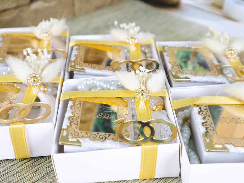 May include: White gift boxes with yellow ribbon and gold accents. Each box contains a small white book with gold lettering, a white tassel, and a string of white beads. The boxes are decorated with white flowers.