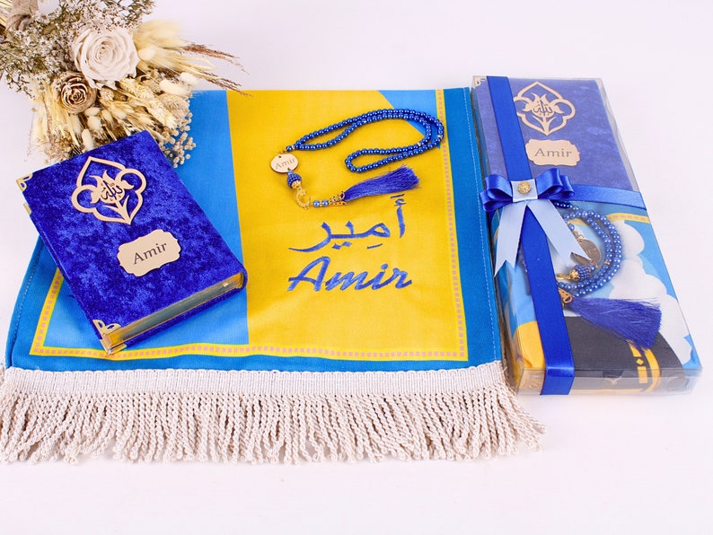 May include: A prayer mat in blue and yellow, featuring the name "Amir" in English and Arabic script, alongside a blue velvet Quran and a blue beaded tasbih. The set is presented with a blue ribbon.
