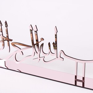 May include: A rose gold mirrored acrylic wall decor with the Arabic phrase "Bismillah" in a decorative font.