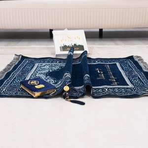 May include: A rolled navy blue prayer mat with a detailed white and silver pattern, fringe, and gold accents. A blue velvet Quran and a gift box with Arabic text are also present. The mat is on a white surface.