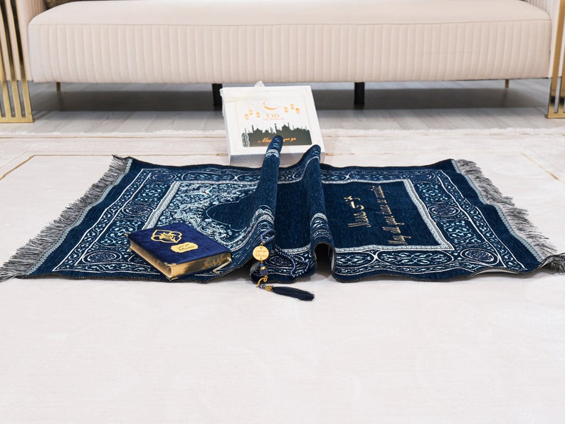 May include: A rolled navy blue prayer mat with a detailed white and silver pattern, fringe, and gold accents. A blue velvet Quran and a gift box with Arabic text are also present. The mat is on a white surface.