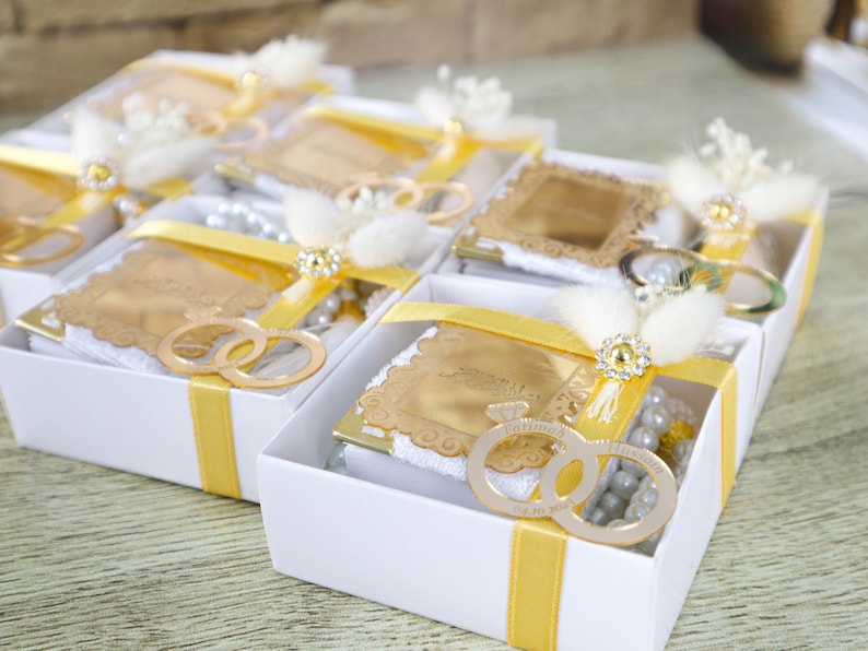May include: White boxes with yellow ribbon tied around them. Each box contains a small gold-colored frame with a white towel inside. The frames have a design of two interlocking rings. The boxes are arranged in a row.