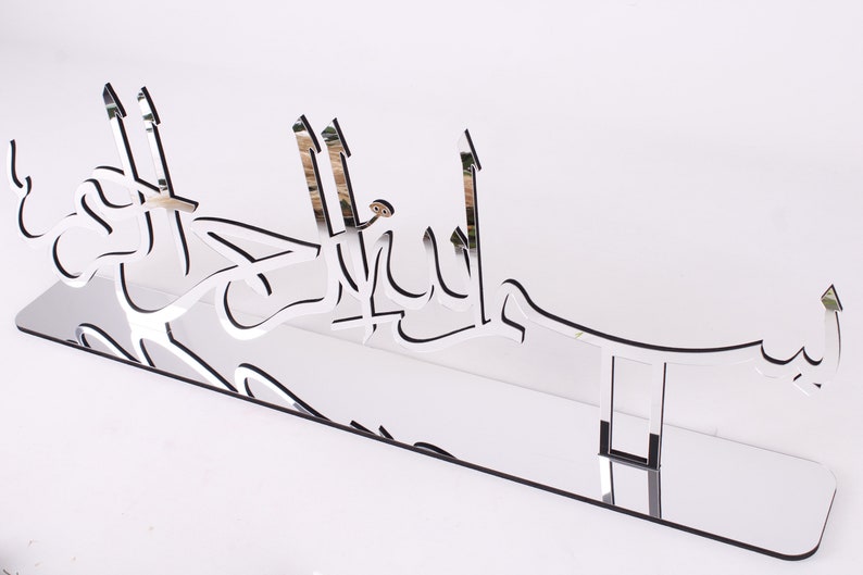 May include: A silver mirrored acrylic wall decor with the Arabic phrase "Bismillah" in a decorative font.