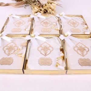 May include: A set of six gift boxes, each containing a white prayer mat, a beaded rosary, and a wooden ornament. The boxes are tied with white ribbons and adorned with gold accents. The boxes are in a gold tray.