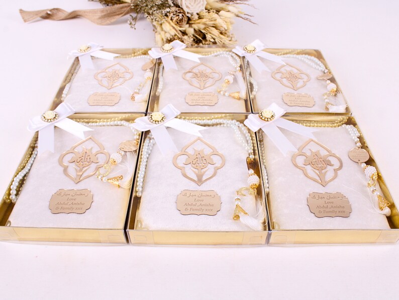 May include: A set of six gift boxes, each containing a white prayer mat, a beaded rosary, and a wooden ornament. The boxes are tied with white ribbons and adorned with gold accents. The boxes are in a gold tray.