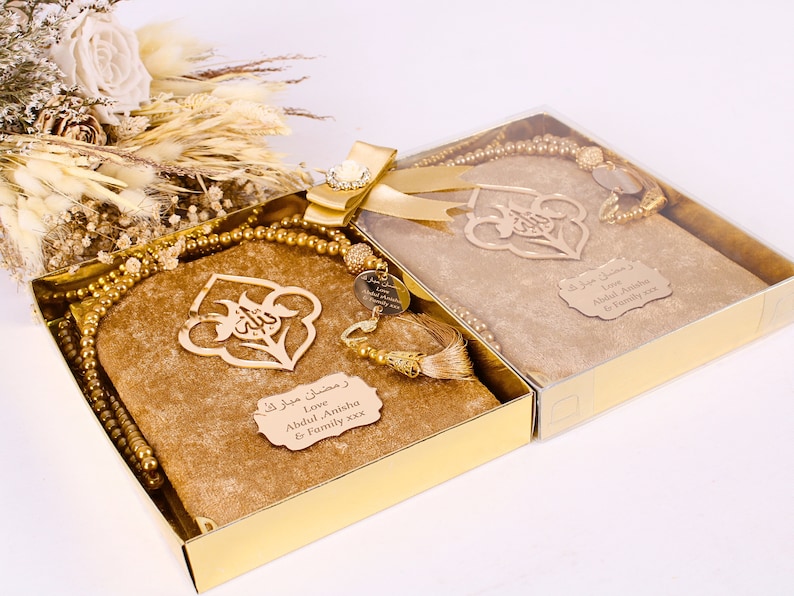 May include: Two gold gift boxes with clear lids, each containing a gold prayer rug, a gold prayer bead necklace, and a gold bookmark with Arabic calligraphy. The boxes are decorated with gold ribbon and a gold jewel. The boxes have personalized messages on the front: "Love Abdul Anisha & Family xxx".