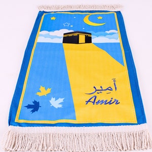 May include: A prayer mat with a blue and yellow design featuring a depiction of the Kaaba, a crescent moon, and stars. The name "Amir" is written in both Arabic and English. The mat has a fringed edge.