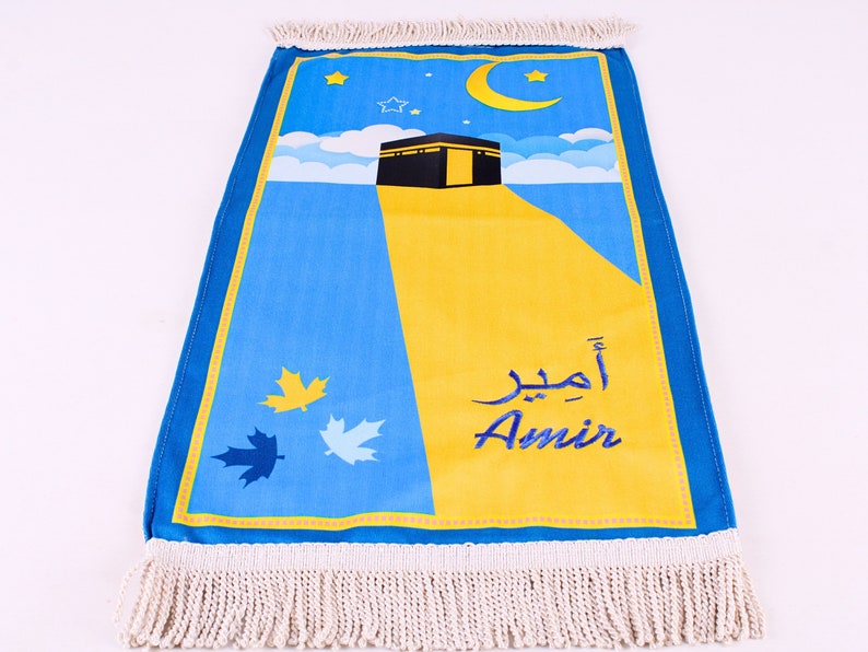 May include: A prayer mat with a blue and yellow design featuring a depiction of the Kaaba, a crescent moon, and stars. The name "Amir" is written in both Arabic and English. The mat has a fringed edge.