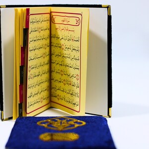K&ouml;nnte beinhalten: Ein kleines, gelbes Buch mit arabischem Text liegt offen auf einem blauen Gebetsteppich. Das Buch hat rote Laschen, die die Seiten markieren. Eine blaue Gebetskette liegt im Vordergrund.