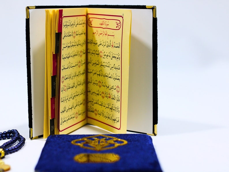 K&ouml;nnte beinhalten: Ein kleines, gelbes Buch mit arabischem Text liegt offen auf einem blauen Gebetsteppich. Das Buch hat rote Laschen, die die Seiten markieren. Eine blaue Gebetskette liegt im Vordergrund.