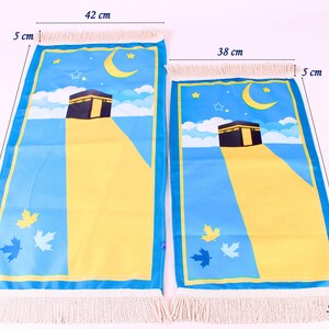 May include: Two prayer mats with a blue and yellow design featuring the Kaaba, a crescent moon, and stars. The larger mat is 88 cm long and 42 cm wide, the smaller one is 68 cm long and 38 cm wide. Both have fringed edges.