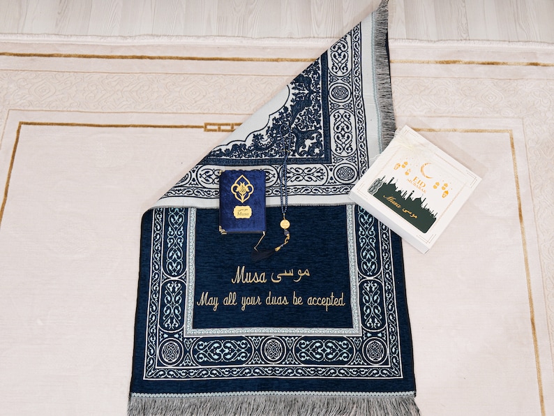 May include: A navy blue prayer mat with white detailing, featuring Arabic script and the English phrase "May all your duas be accepted." Includes a small blue book, prayer beads, and a gift box with "Eid Mubarak" text.