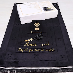 May include: A black prayer rug with gold embroidery reading "Amir" and "May all your duas be accepted," featuring a black book with a gold emblem and a black beaded tasbih. Two white gift boxes with gold trim are placed at the top of the rug.