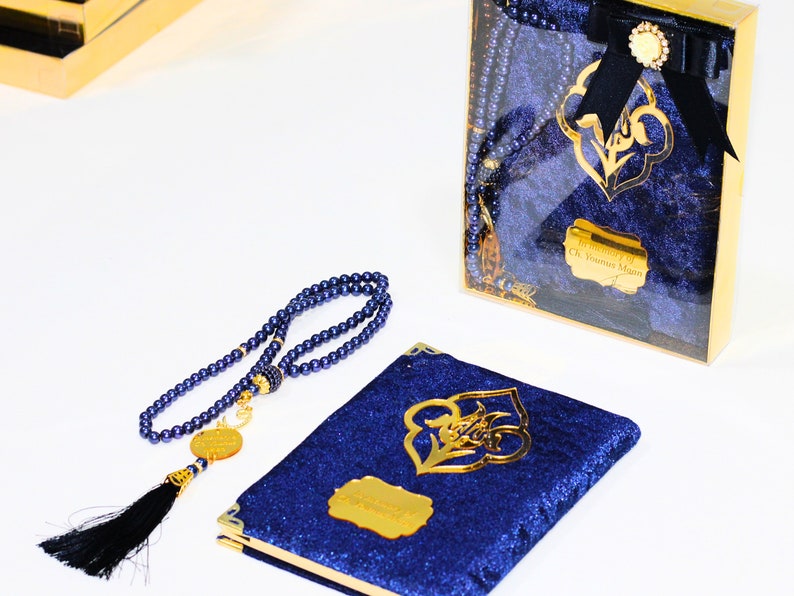K&ouml;nnte beinhalten: Ein Gebetbuch aus blauem Samt mit goldenem Design und einer passenden blauen Gebetskette mit goldener Quaste. Das Buch befindet sich in einer durchsichtigen Schachtel mit goldener Einfassung und dem Text "In memory of Ch. Yousuf Mian".