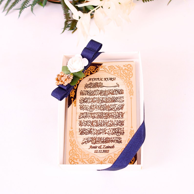 May include: A wooden plaque with gold lettering and a blue ribbon. The plaque features the Arabic text of Ayatul Kursi, a verse from the Quran, and is personalized with the names "Amir & Zainab" and the date "12.12.2022".