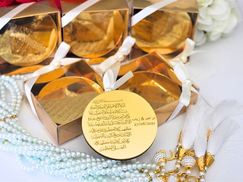 Op de afbeelding: Gouden geschenkdozen met wit lint en een ronde gouden plaquette met Arabische kalligrafie en de datum 27/06/2021. De plaquette is omgeven door witte parels en witte kwastjes.