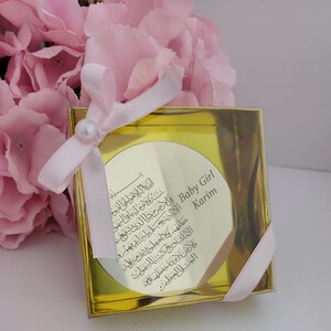 Op de afbeelding: Een gouden geschenkdoos met een roze lint en een parel. De doos bevat een ronde, gouden plaquette met Arabische kalligrafie en de tekst "Baby Girl Karim".