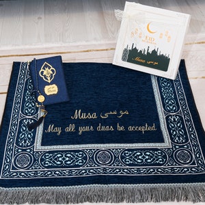May include: A navy blue prayer mat with intricate white and teal patterns, alongside a matching Quran cover. The mat features gold embroidery of the name "Musa" and the phrase "May all your duas be accepted." A gift box with "Eid Mubarak" is also visible.
