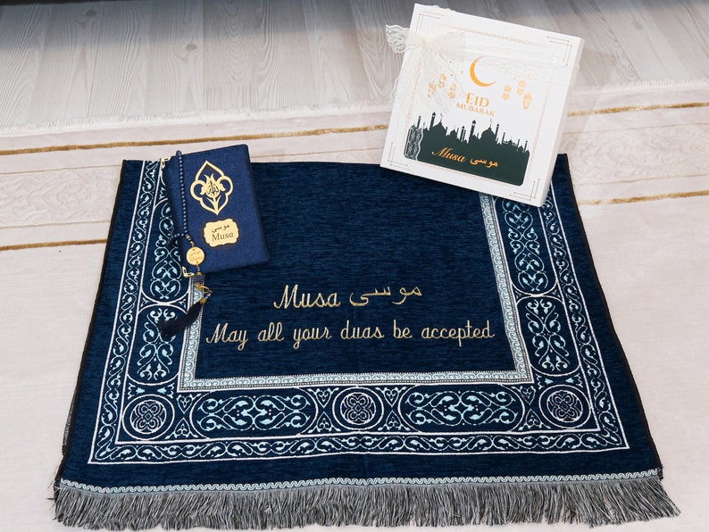 May include: A navy blue prayer mat with intricate white and teal patterns, alongside a matching Quran cover. The mat features gold embroidery of the name "Musa" and the phrase "May all your duas be accepted." A gift box with "Eid Mubarak" is also visible.