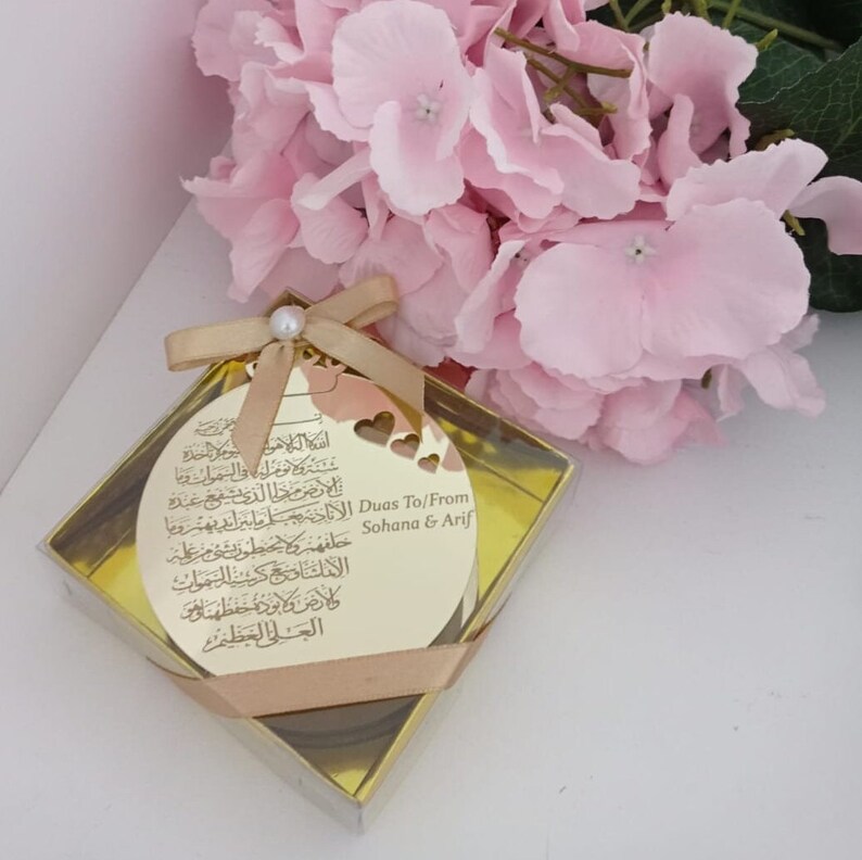 Op de afbeelding: Een gouden ronde ornament met Arabische kalligrafie en een hartvormige uitsparing. De ornament bevindt zich in een doorzichtige doos met een gouden lint en een parel accent. De tekst op de ornament luidt "Duas To/From Sohana & Arif".