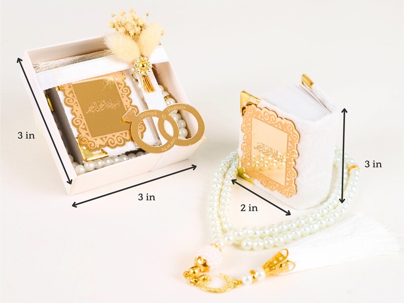 May include: A white gift box with a white book inside, a white prayer beads necklace, and two gold rings with Arabic calligraphy. The box is 3 inches wide and 3 inches tall. The necklace is 3 inches long and 2 inches wide.