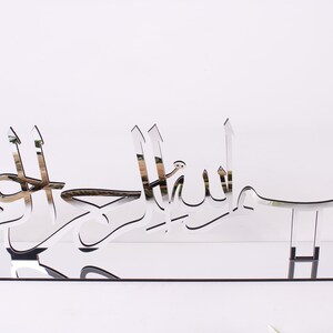 May include: A silver mirrored wall decor with the Arabic phrase "Bismillah Al Rahman Al Rahim" which translates to "In the name of Allah, the Most Gracious, the Most Merciful".