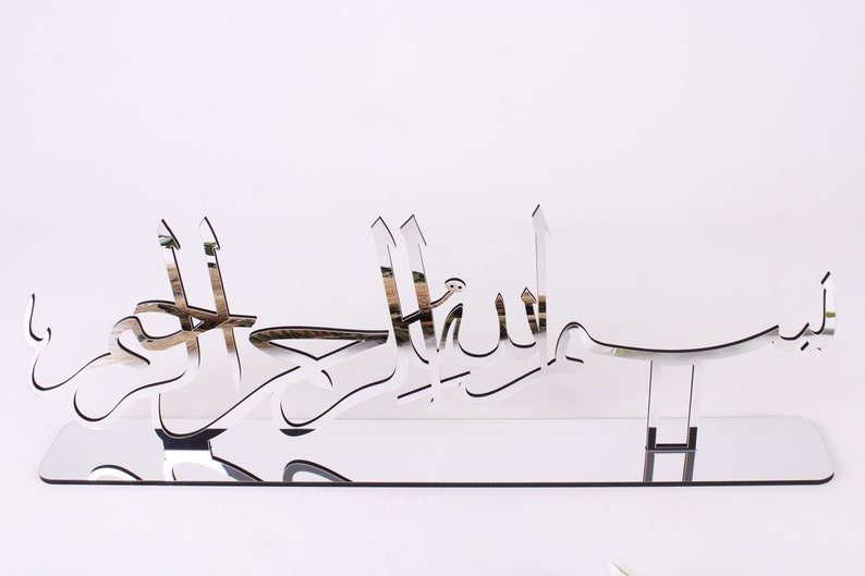 May include: A silver mirrored wall decor with the Arabic phrase "Bismillah Al Rahman Al Rahim" which translates to "In the name of Allah, the Most Gracious, the Most Merciful".