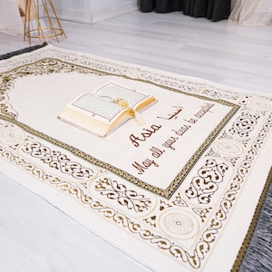 Puede incluir: Una alfombra de oración blanca con un borde dorado y negro. La alfombra tiene una inscripción personalizada que dice "Asia Que todas tus súplicas sean aceptadas". Un Corán está colocado sobre la alfombra.