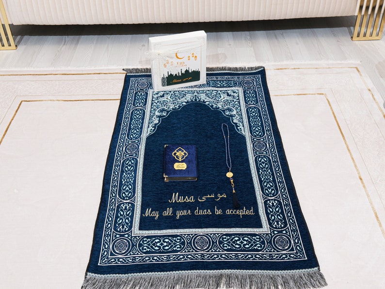 May include: A navy blue prayer mat with an arched design and detailed white and light blue patterns. A small blue book with gold accents and a string of prayer beads are on the mat. The text "Musa" and "May all your duas be accepted" are embroidered.