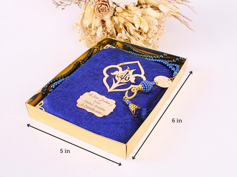 May include: A gold box containing a blue prayer mat, a beaded rosary, and a decorative gold pendant. The box is 5 inches wide and 6 inches long. The box also contains a small gold plaque with Arabic text.