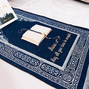 Puede incluir: Una alfombra de oración azul marino con patrones blancos y un borde decorativo. Un libro abierto y cuentas de oración descansan sobre la alfombra. El texto "Musa" y "May all your duas be accepted" están bordados en oro.