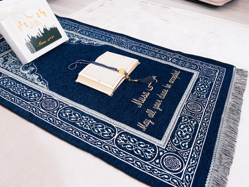 May include: A dark blue prayer mat with white decorative patterns and a border. An open book and prayer beads are on the mat. The text "Musa" and "May all your duas be accepted" are embroidered in gold.