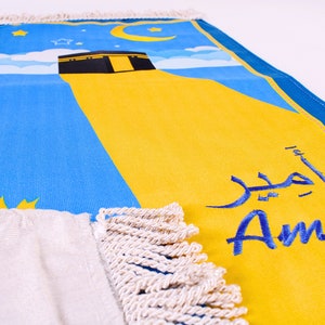 May include: A prayer mat featuring a blue sky design, a yellow section, and a black structure. The mat has the name "Amir" in Arabic and English. The mat has a white fringe.