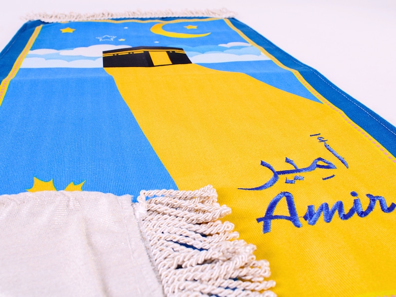 May include: A prayer mat featuring a blue sky design, a yellow section, and a black structure. The mat has the name "Amir" in Arabic and English. The mat has a white fringe.