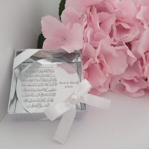 Op de afbeelding: Een zilveren doosje met een wit lint en een ronde, doorzichtige schijf erin. De schijf heeft Arabische kalligrafie en de tekst "Sara & Sherif 9/5/21".
